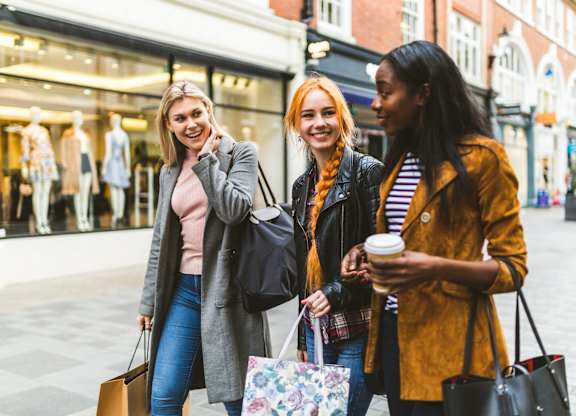 Three women are walking down the street with shopping bags and coffee.