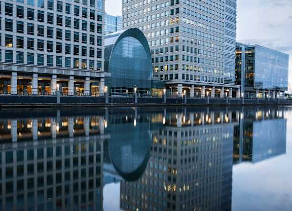 A modern building with a curved glass facade is reflected in the water.