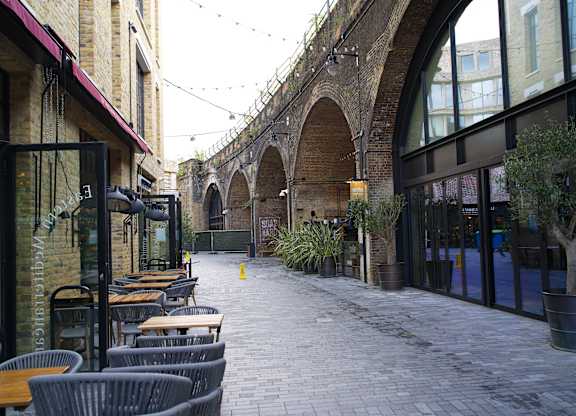 A long alley with tables and chairs on the left and a building with a glass door on the right.