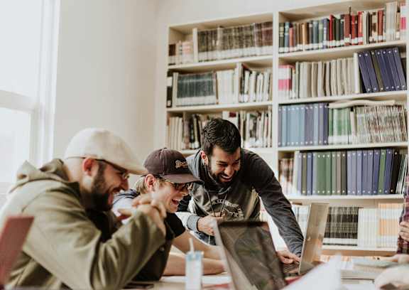 A group of people are gathered around a laptop in a room with bookshelves in the background.