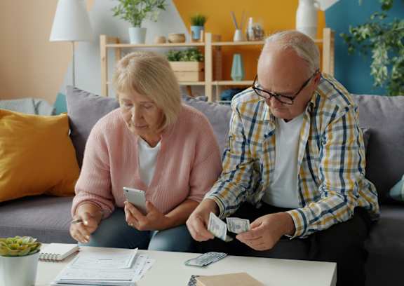 A man and a woman are sitting on a couch, looking at a smartphone and counting bills.