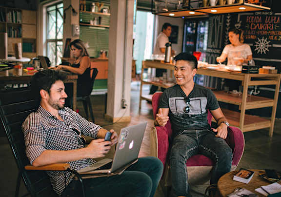 Two men are sitting in a cafe, one is using a laptop and the other is holding a phone.