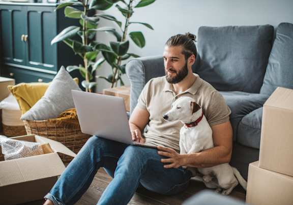 A man is sitting on the floor with a laptop, surrounded by cardboard boxes and a dog is resting on his lap.