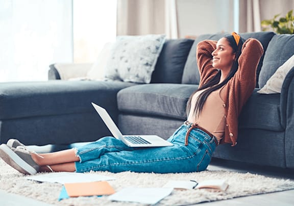 A woman is sitting on the floor with her laptop and her feet up on a couch.