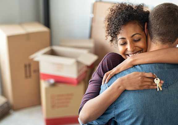 A couple hugging each other in a room with moving boxes.