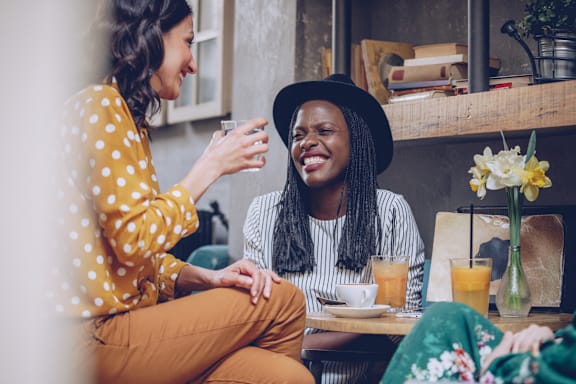 Two women are sitting at a table, one is holding a glass and they both appear to be laughing.