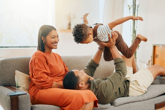 A family of three is sitting on a grey couch, with the child on the man's shoulders.