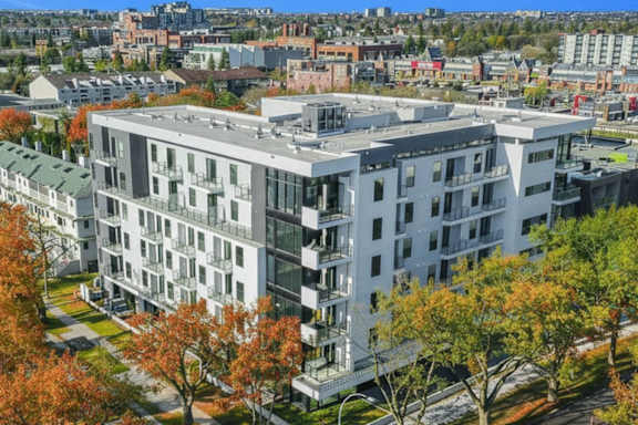 A modern multi-story building with balconies is surrounded by trees with autumn leaves.