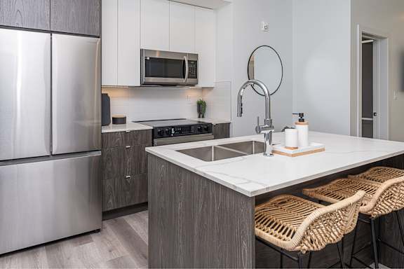 A modern kitchen with a stainless steel refrigerator, a microwave above the stove, and a white countertop with two wicker chairs.