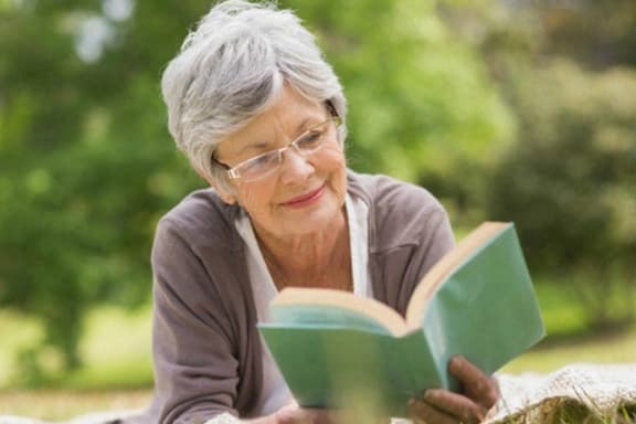 an older woman with glasses reading a book in a park