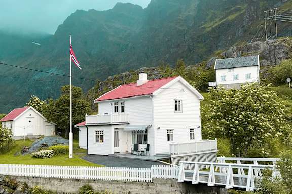 A white house with a red roof is situated by a body of water with a bridge leading to it.
