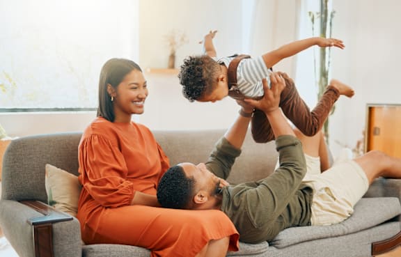 A family of three is sitting on a grey couch, with the child on the man's shoulders.