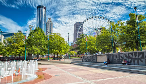 a city square with a fountain in the foreground and a ferris wheel in the background