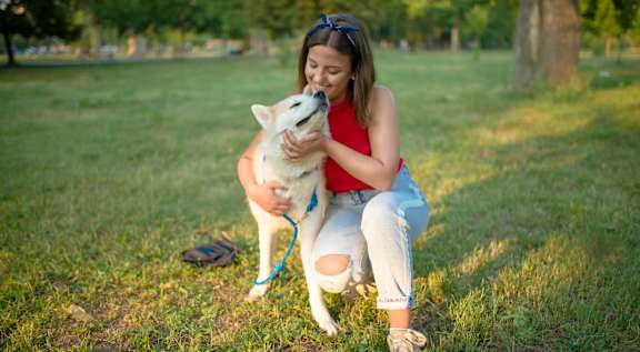 A woman in a red top and ripped jeans is sitting on the grass with a white dog.