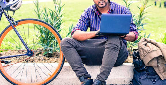 A man is sitting on the ground with a laptop and a bicycle beside him.