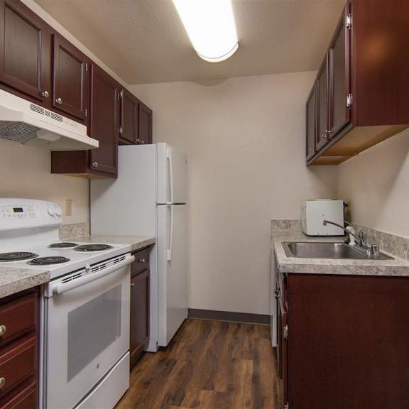 compact kitchen here at outlook apartments with dark cherry cabinets, white appliances, and a stone-patterned countertop. wooden floor adds warmth. bright, clean, and modern.