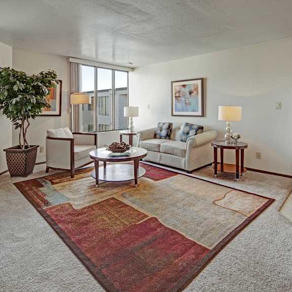 a bright living room at the alpine featuring a sofa and armchair arranged around a round coffee table on a colorful area rug. large windows bring in natural light and views of the outdoors. table lamps, framed artwork, and a potted plant add warmth, with the kitchen area visible nearby.