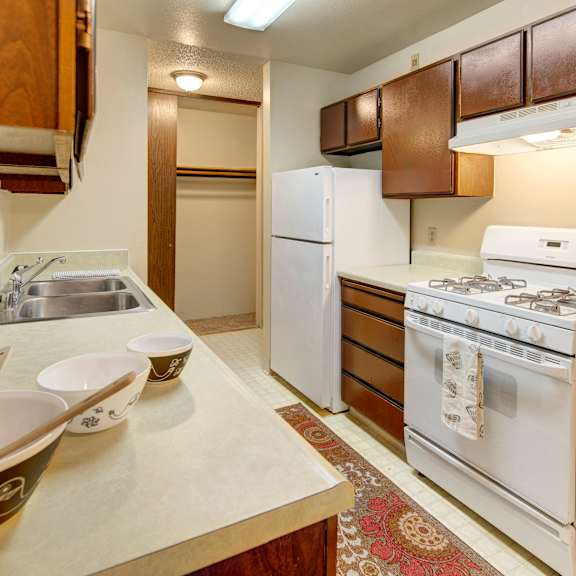 narrow kitchen here at taiga apartments with brown cabinets, white appliances including a refrigerator and gas stove, three bowls on the counter, and a patterned rug on the floor.