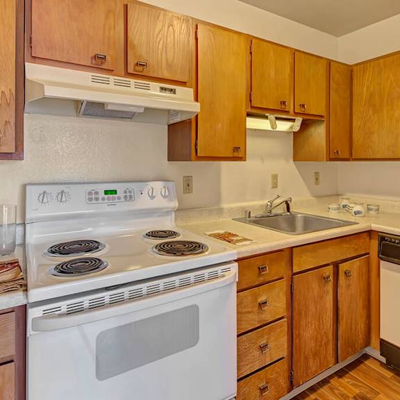 cozy kitchen with wooden cabinets, white electric stove, and sink with overhead lights. counters have a few decor items, creating a warm atmosphere. the alaskan apartments.