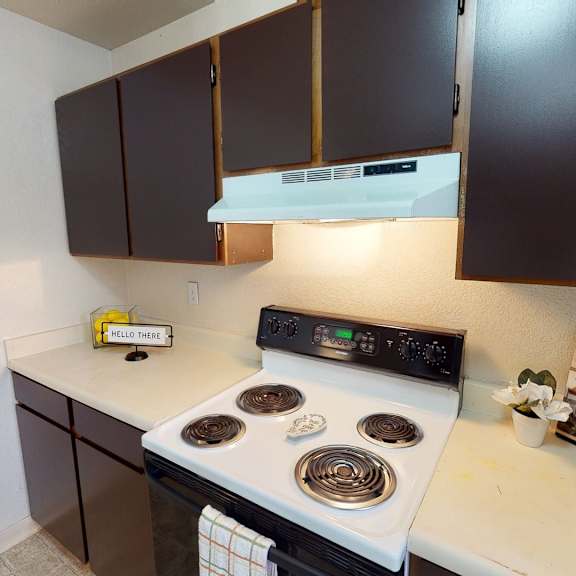 cozy kitchen here at deer park apartments with dark brown cabinets, white stove, and beige countertops. cheerful decor includes a "hello there" sign and a small plant.