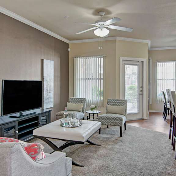 a bright and inviting living room here at mozaic at steele park featuring plush carpet, neutral walls, and a ceiling fan with light. large windows with vertical blinds and a glass door bring in natural light, while the open layout flows into a kitchen with bar seating, creating a comfortable space for relaxing or entertaining.