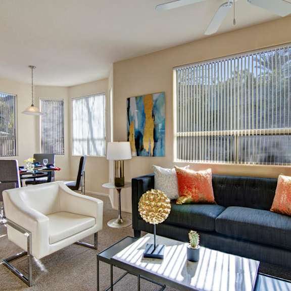 a welcoming living room here at sonoma landing featuring a dark upholstered sofa accented with warm orange pillows, a sleek glass coffee table with modern décor, and a white accent chair, while large windows with vertical blinds fill the space with natural light and connect to a dining area with a contemporary table, chairs, and pendant lighting.