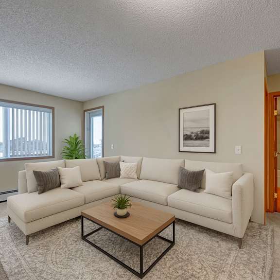 a bright, modern living room here at sonora featuring a spacious neutral sectional sofa with accent pillows, a wooden coffee table on a soft area rug, large windows with vertical blinds bringing in natural light, and a clean, open layout connecting comfortably to the adjacent hallway and bathroom.