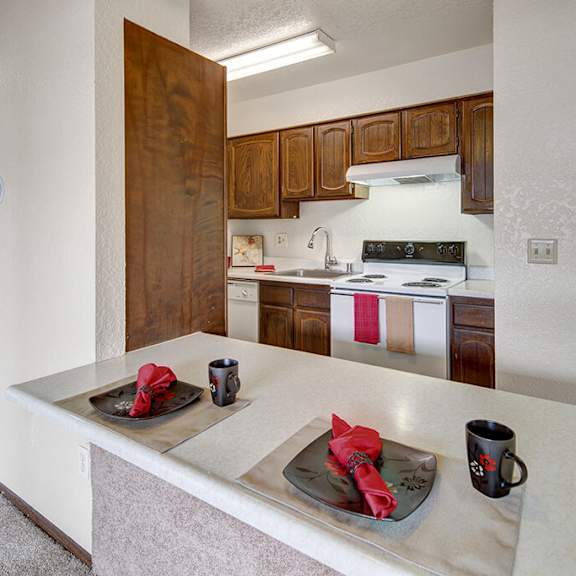 a cozy kitchen here at the continental apartments with wooden cabinets and white appliances. a countertop features two place settings with black dishes, red napkins, and mugs, creating a warm, inviting atmosphere.