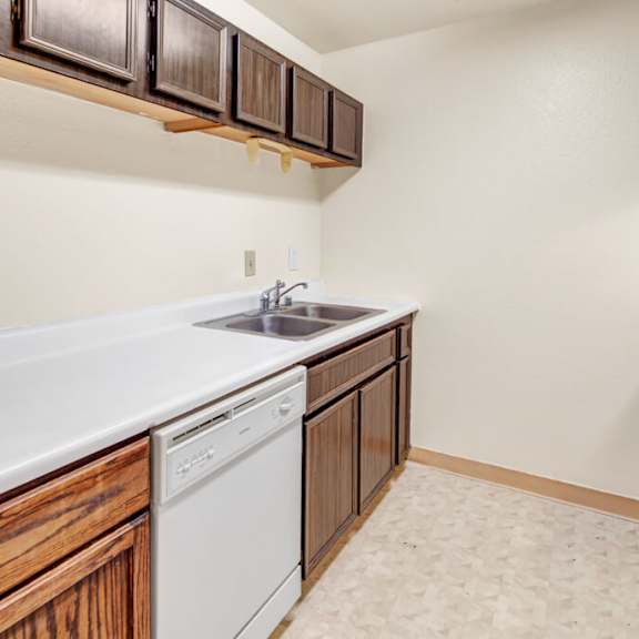 a narrow kitchen here at ladera villas apartments with dark brown cabinets, white countertops, and a stainless steel sink. a dishwasher is below, and the room has a neutral, minimalist tone.