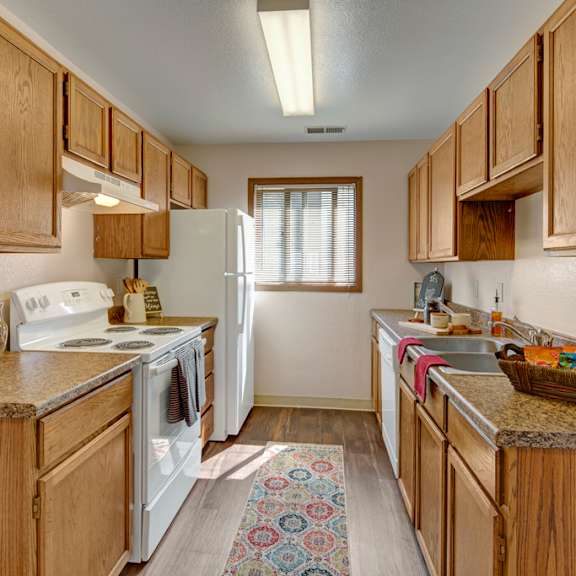narrow kitchen here at legacy west apartments with wood cabinets, white appliances, and granite countertops. a rug adds warmth, while natural light streams through a window. cozy ambiance.