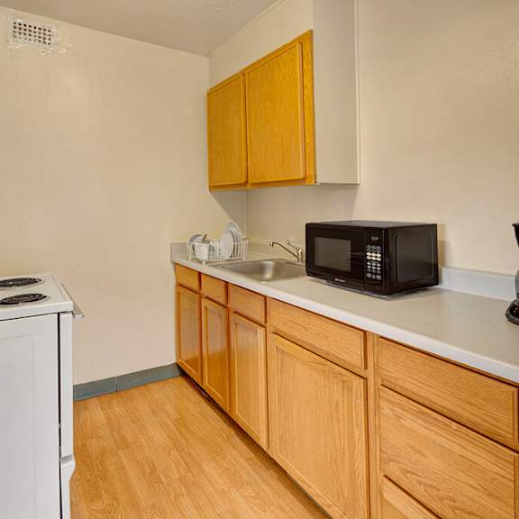 compact kitchen here at mendenhall tower apartments with light wood cabinets, white appliances, and a microwave. includes an electric stovetop, sink with dish rack, and a coffee maker.