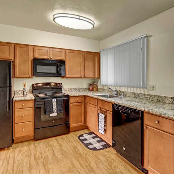 a modern kitchen here at monctlair apartments with wooden cabinets, black appliances including a fridge, stove, and dishwasher, granite countertops, and a window with vertical blinds.