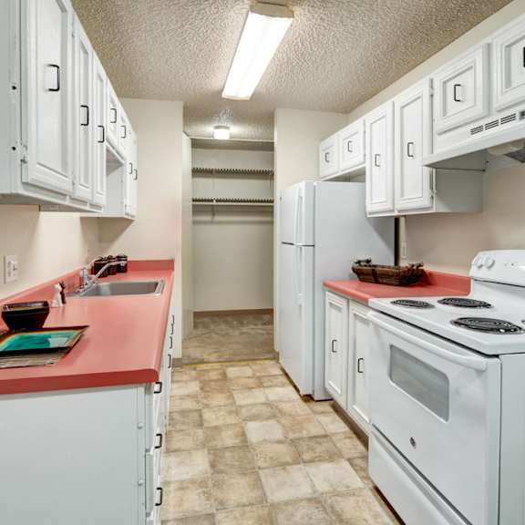 compact kitchen here at quiet creek apartments with a retro feel features white cabinets, a red countertop, and white appliances. bright lighting highlights the clean, orderly space.