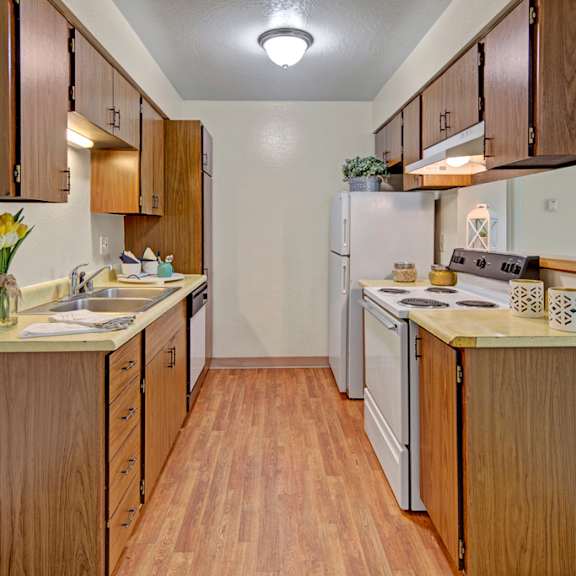 a narrow kitchen here at rancho tudor apartments with wood cabinets and laminate flooring. yellow flowers in a vase are on the counter, alongside neatly arranged kitchenware, creating a warm, welcoming ambiance.