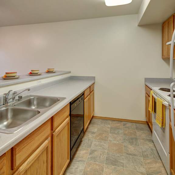 modern kitchen here at strawberry lane apartments with light wood cabinets, dual sink, white appliances, and gray countertops. bright with orange decor and tiled floor, creating a cozy feel.