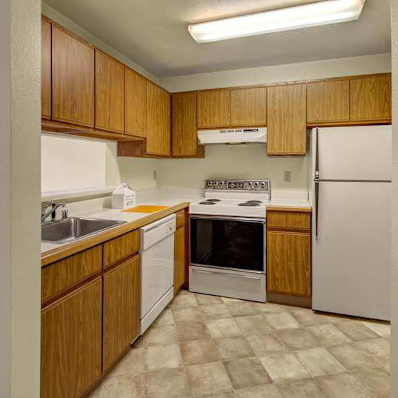 compact kitchen here at susitna ridge apartments with wooden cabinets, white appliances including a stove, refrigerator, dishwasher, and a sink. tiled floor and bright lighting.