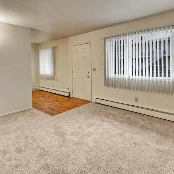 a vacant living room here at village at calais apartments with light beige walls and carpet, featuring a large window with vertical blinds. the entry area has hardwood flooring.