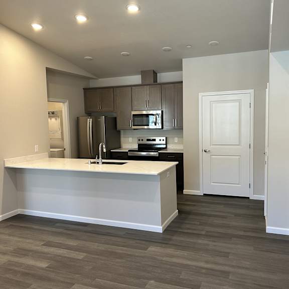 a contemporary open living room here at autumn haus featuring wide plank flooring, recessed ceiling lights, and an expansive kitchen with a large quartz-style island, undermount sink, modern cabinetry, and sleek appliances, creating a bright, functional layout that flows seamlessly into the main living space.