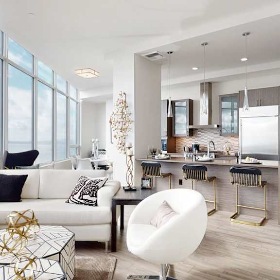 a bright, modern living room here at tower 12 with floor-to-ceiling windows and water views, wood flooring, and an open kitchen featuring bar seating, stainless steel appliances, and in-unit laundry nearby.