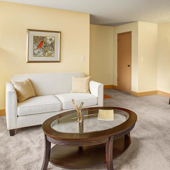 a cozy living room here at whispering cedars featuring a light beige sofa with neutral pillows, round glass coffee table, warm floor lamp, soft carpet, and a simple desk area creating a comfortable, inviting space.