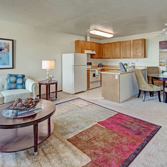 a cozy living and kitchen area with beige walls and carpet. a white sofa hosts patterned cushions beside a round table with a lamp. the kitchen features wood cabinets, and a dining area with a round table and tan chairs. a colorful abstract rug and artwork add warmth. located at alpine apartments.