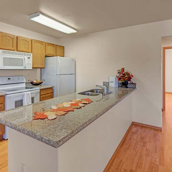 modern kitchen with light wood cabinets, stone countertop, white appliances, and fall-themed decorations. bright lighting and wood flooring add warmth. amber ridge apartments.