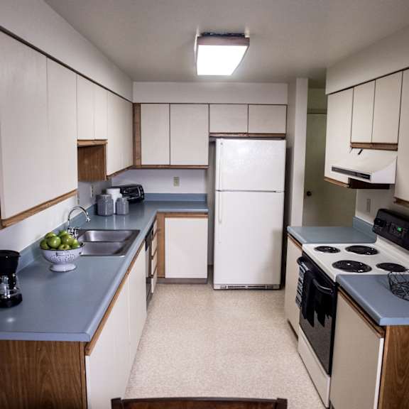 a narrow kitchen here at birchwood apartments with white cabinets, blue countertops, a refrigerator, stove, and sink. a bowl of green apples and a coffee maker are visible.