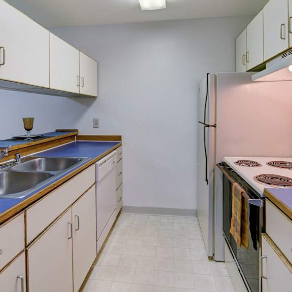 small kitchen here at chugach south apartments with blue countertops, white cabinets, and a four-burner stove. includes a double sink, fridge, and cozy setting with warm lighting.