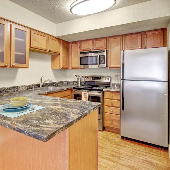 compact kitchen here at city view apartments with light wood cabinets and marble countertops. stainless steel appliances, including a fridge and microwave. cozy and inviting feel.