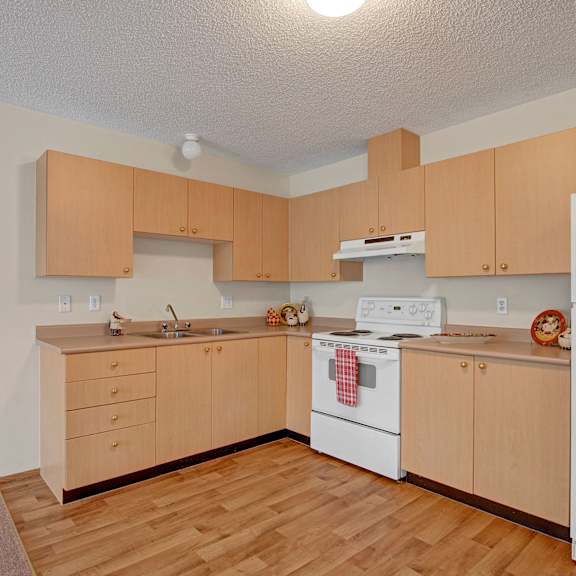 compact kitchen here at birch terrace apartment homes with light wood cabinets, white appliances including a fridge and stove, double sink, and wooden floor, conveying a clean and cozy feel.