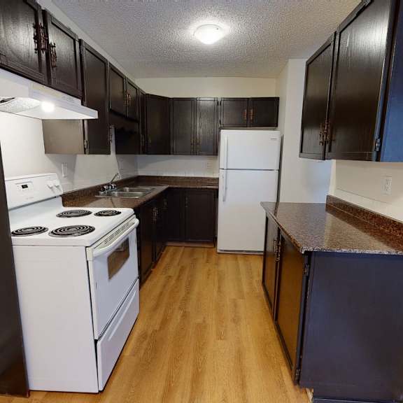 narrow kitchen here at broadmoor apartment homes with dark wood cabinets, white appliances, and a light wood floor. bright, inviting atmosphere with ample counter space and overhead lighting.