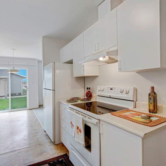 compact kitchen here at carrington place apartment homes with white cabinets, stove, and fridge. sunlit space via glass door to yard. cozy, clean ambiance with minimal decor.