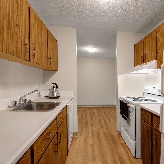 narrow kitchen here at gateway gardens apartment homes with wooden cabinets, white countertops, stainless steel sink, and kettle. white appliances include a fridge and stove. cozy and simple.