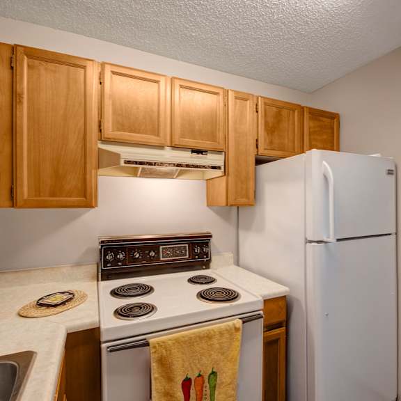 compact kitchen here at gateway square apartment homes with wooden cabinets, electric stove, and white fridge. a towel with pepper designs hangs on the oven handle, creating a cozy vibe.